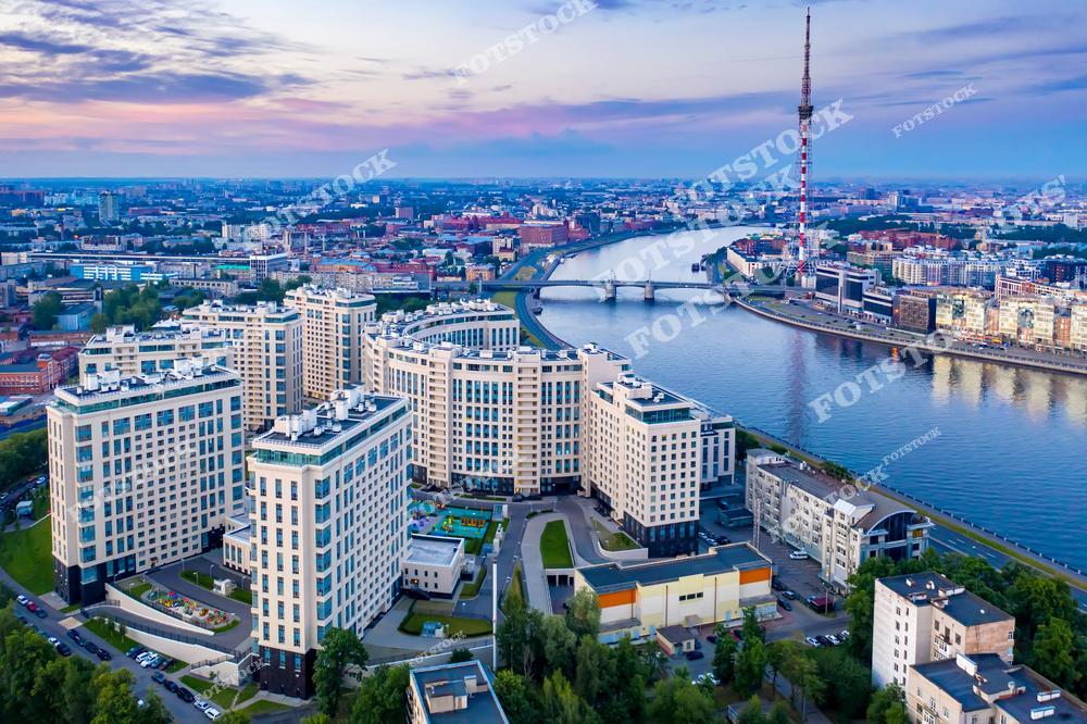 На фотографии запечатлена атмосфера белых ночей в Санкт-Петербурге. Современные жилые комплексы на берегу Невы сочетаются с городской инфраструктурой. Воды реки отражают ночные огни, создавая живописный пейзаж. Это уникальное время года подчёркивает романтику и динамику северной столицы.